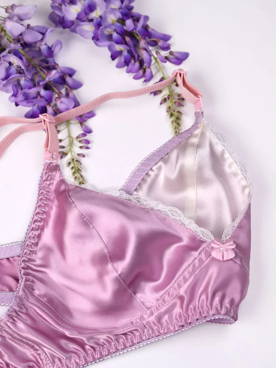 Pink satin bralette with lace details on a white background with purple flowers.